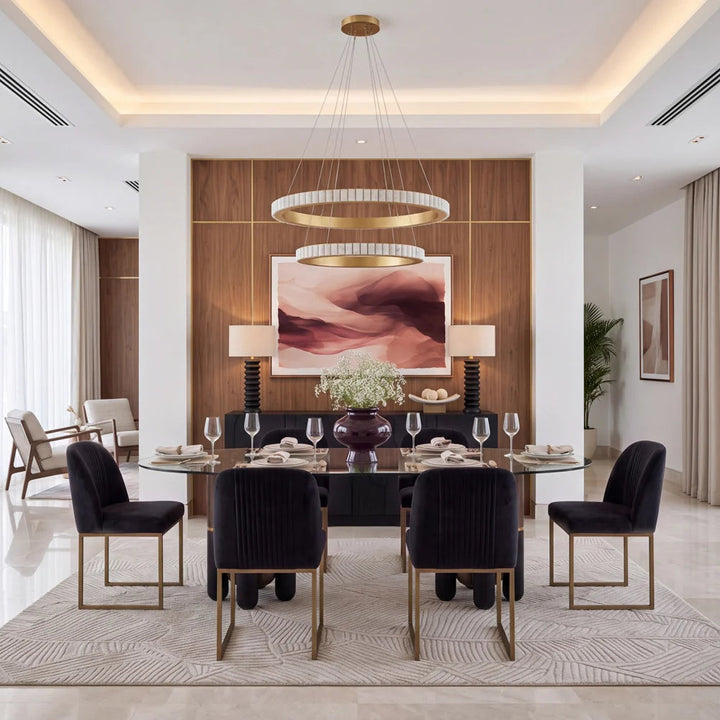 A luxury modern dining room featuring a round glass-top dining table with six dark velvet dining chairs with gold legs, set on a patterned area rug. The background features a warm wood-paneled wall with a large abstract art print, flanked by two black side tables with white lamps.