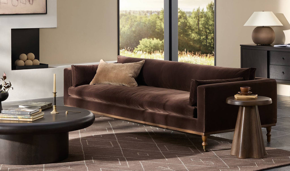 A cocoa velvet sofa with sculptural tables and a ceramic lamp in a sunlit living room
