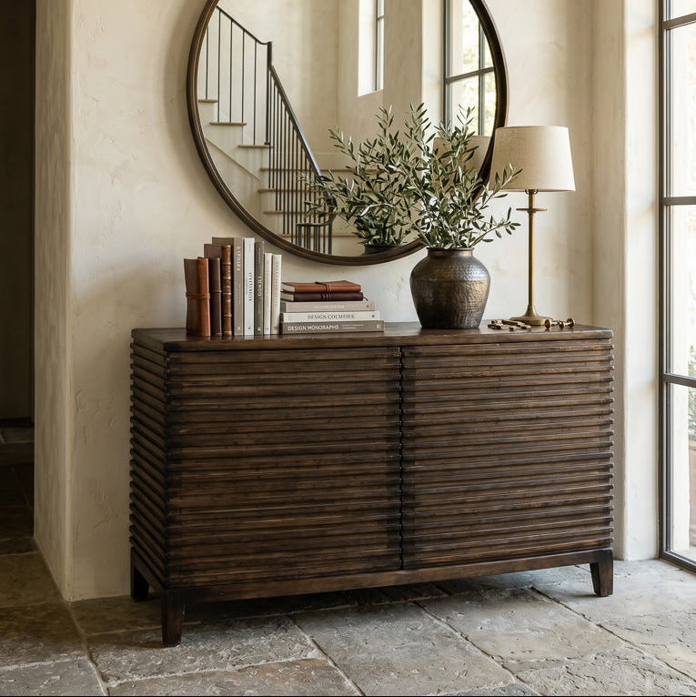 A dark brown wooden console cabinet with a slatted front, styled in a home entryway under a large round mirror. The cabinet is decorated with books, a vase of greenery, and a table lamp, set against a textured beige plaster wall.