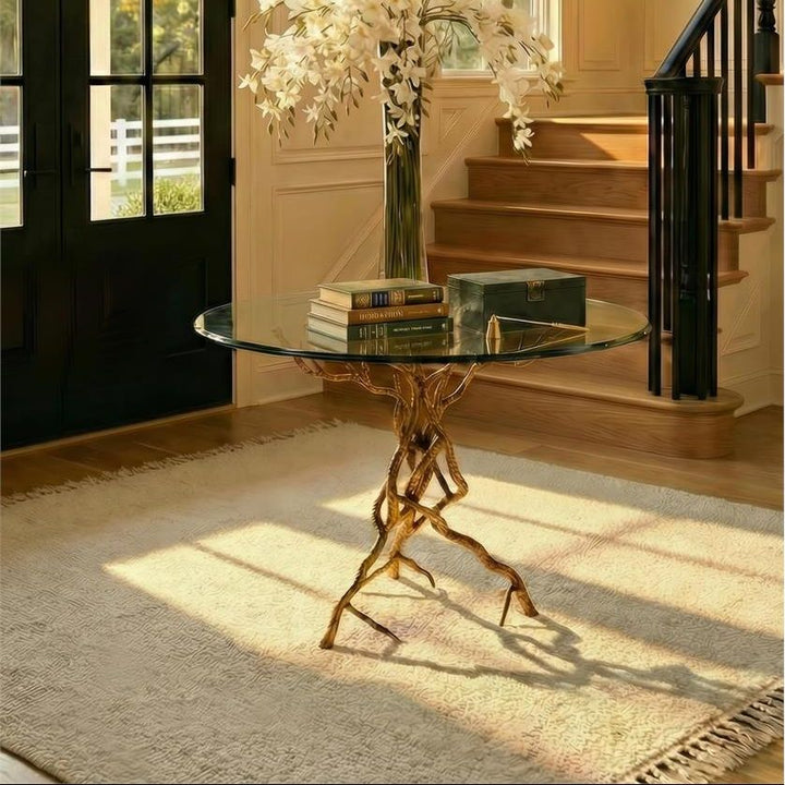A grand entryway with a round glass-top console table featuring a sculptural gold base, styled with a tall vase of white flowers and books. The room has cream walls, a staircase with a black railing, and a large chandelier overhead.