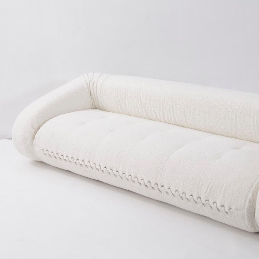 A white fabric-covered curved sofa cushion or small sofa shown in a plain indoor setting with a white floor and wall, plus a blue crate partially visible in the background.