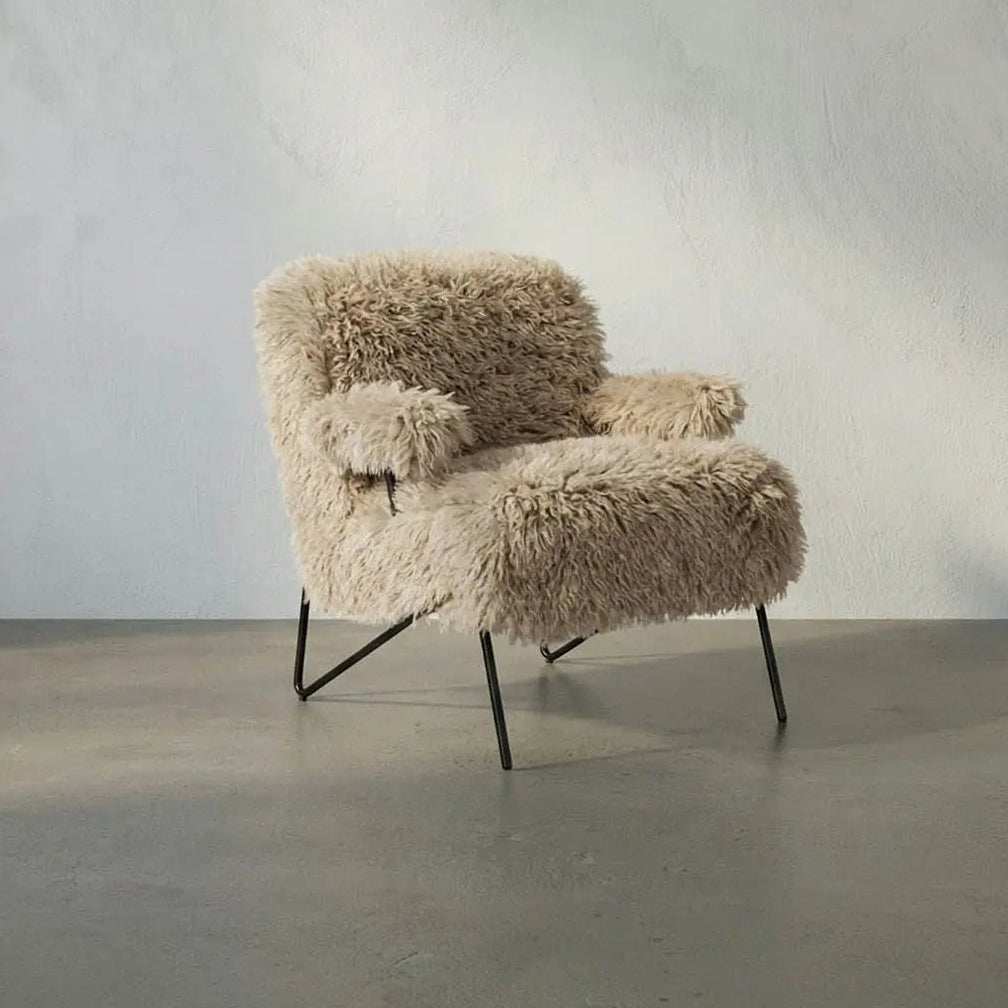 A modern low-profile armchair upholstered in shaggy beige faux fur or sheepskin, supported by slim black metal legs, set against a textured light gray plaster wall and concrete floor.