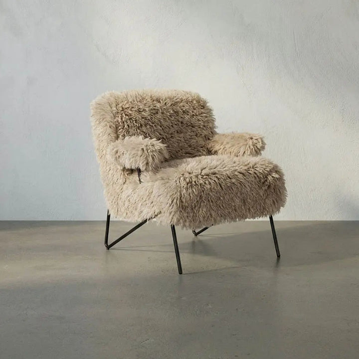 A modern low-profile armchair upholstered in shaggy beige faux fur or sheepskin, supported by slim black metal legs, set against a textured light gray plaster wall and concrete floor.