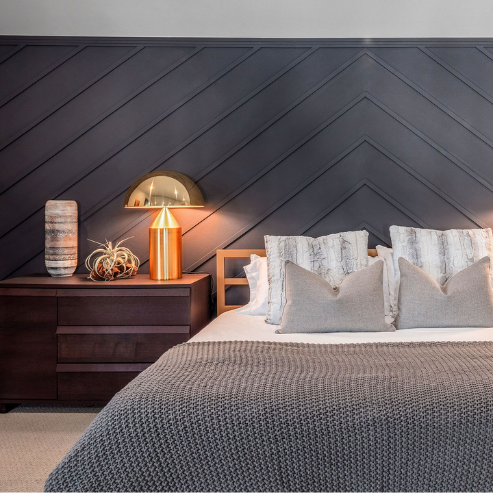A modern bedroom setup featuring a dark wood chevron-paneled headboard wall, matching dark wood nightstands, a textured gray bedspread, and two matching copper table lamps with gold shades on either side of the bed.