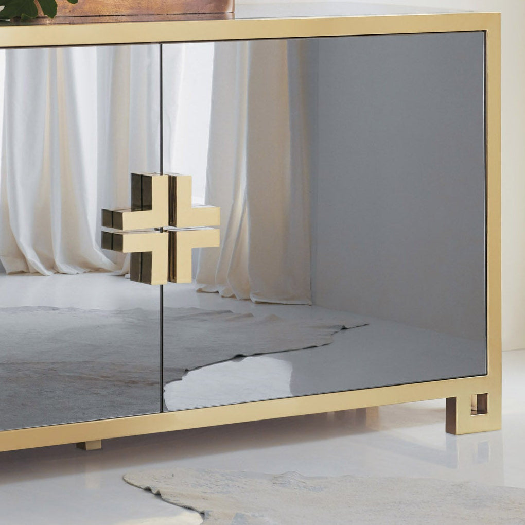 A modern console table with a gold metal frame, mirrored glass doors, and gray side panels, styled in a minimalist room setting with a small potted plant on top.