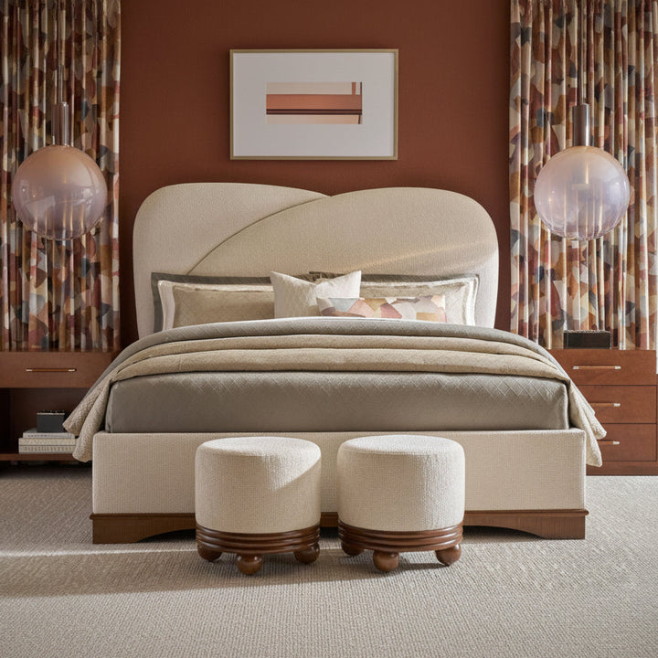 A styled bedroom scene featuring a cream upholstered bed with a sculptural curved headboard, matching bed skirt, patterned beige and gray bedding, two matching cream ottomans at the foot of the bed, and patterned curtains against a warm brown wall.