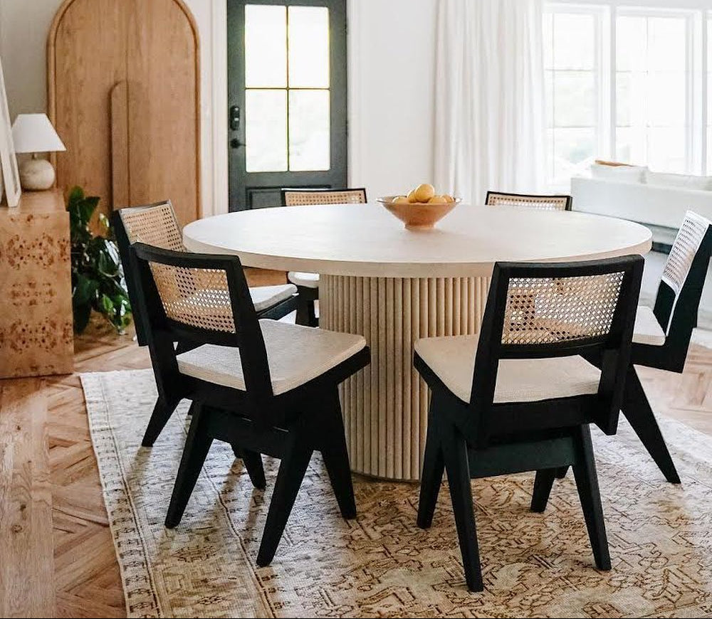 Dining room with round table and black chairs with white cushions, featuring a modern chandelier.